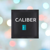 A black package labeled "Caliber" with a small turquoise design is centered against a blurred pastel lights background, showcasing the Caliber M - Standard Size Condoms (53mm Wide).